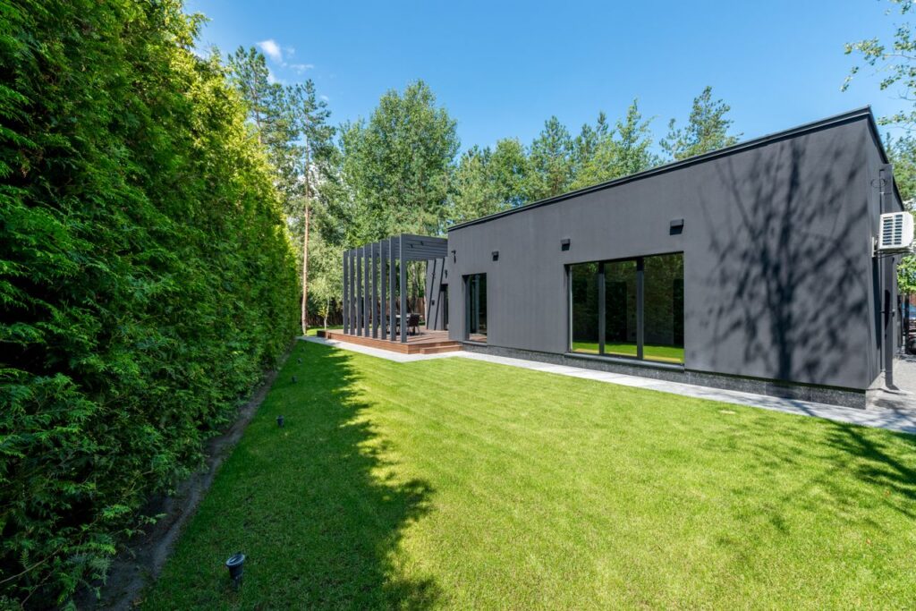 tonte pelouse Sleek black modern house with large windows and vibrant green lawn surrounded by dense trees.